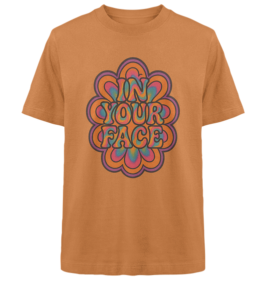Oranges T-Shirt mit großem, psychedelischen Retro-Blumenmuster in kräftigen Regenbogenfarben. In der Mitte steht der Satz „IN YOUR FACE“ in verschnörkelter 70er-Jahre-Typografie, umrandet von einer schwarzen Outline. Das Design wirkt auffällig, bunt und provokant.